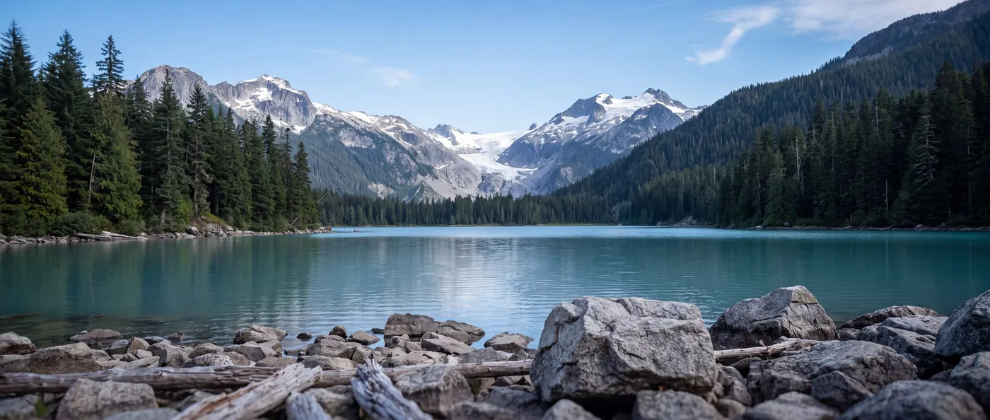 A glacial lake in the Coast Mountains of British Columbia, surrounded by conifer forest, with snow-capped peaks above it